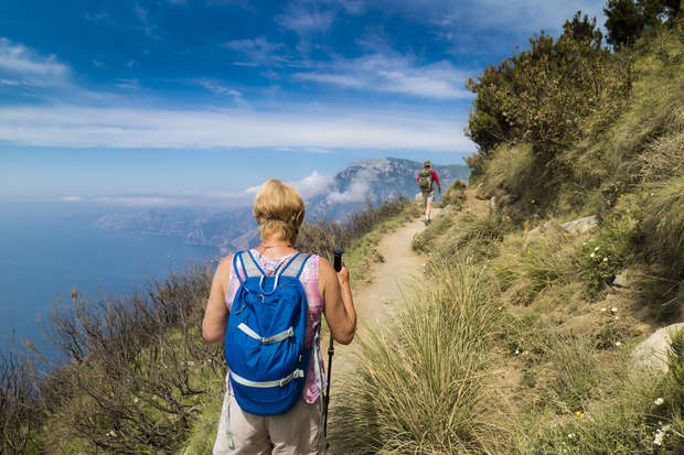 A person with a blue backpack walks along a scenic mountain trail, surrounded by grass, bushes, and overlooking a distant ocean under a clear blue sky.